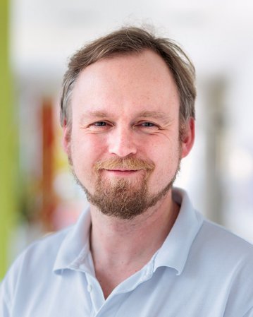 Ein Mann mit einem kurzen Bart und lässiger Kleidung lächelt in die Kamera, vor einem unscharfen Hintergrund.