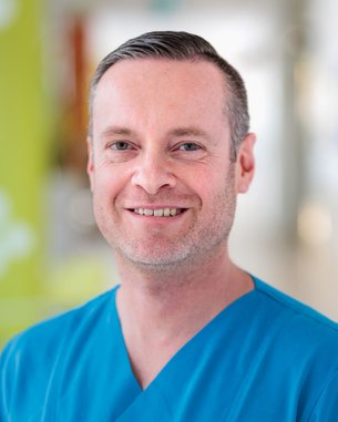 Ein Mann in einem blauen medizinischen Outfit lächelt freundlich in die Kamera, mit einem hellen, unscharfen Hintergrund.