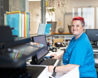 Eine lächelnde Frau mit kurzen, bunten Haaren trägt ein blaues Oberteil und sitzt an einem Schreibtisch mit einem Computer. Im Hintergrund sind farbige Ordner und ein Drucker sichtbar.