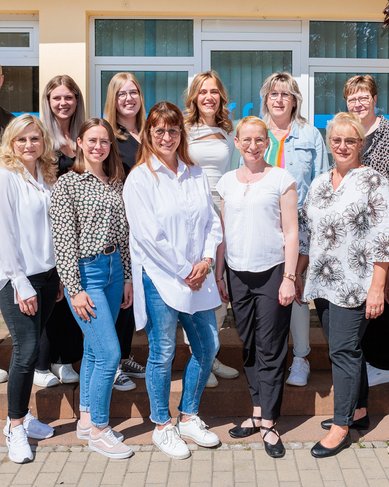 Gruppenfoto von 13 Personen, die auf einer Treppe stehen. Die Gruppe besteht aus Frauen und einem Mann, die in verschiedenen Outfits gekleidet sind. Im Hintergrund sind Fenster und ein Teil des Gebäudes sichtbar.