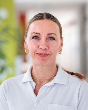 Eine Frau mit langen, blonden Haaren trägt ein weißes Polohemd und goldene Ohrringe. Sie steht in einem hellen, modernen Raum.