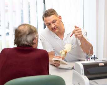 Ein Arzt erklärt einem älteren Patienten mit Hilfe eines anatomischen Modells das Gelenk. Der Arzt zeigt mit einem Stift auf das Modell, während der Patient aufmerksam zuhört.