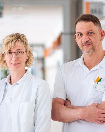 Drei medizinische Fachkräfte in weißen Kitteln stehen in einem Flur eines Krankenhauses. Sie lächeln und wirken professionell.