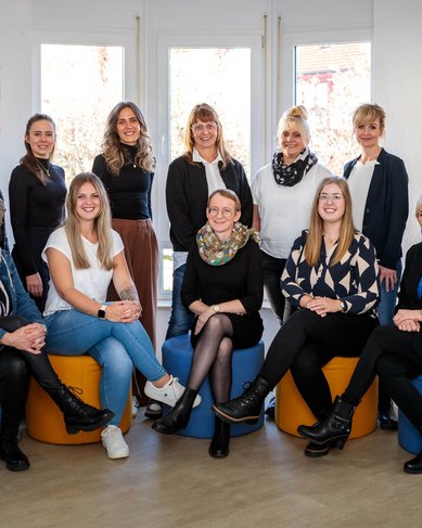 Eine Gruppe von zwölf Frauen sitzt auf farbigen Hockern in einem hellen Raum. Sie tragen verschiedene formelle Outfits und lächeln in die Kamera.
