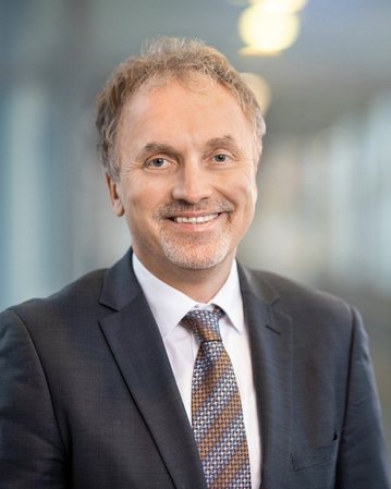 Ein lächelnder Mann mit kurzen, grauen Haaren trägt einen Anzug und eine Krawatte. Der Hintergrund zeigt unscharfe, helle Lichter und eine moderne Umgebung.