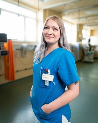 Eine Krankenschwester in blauer Uniform steht in einem Krankenhauszimmer und lächelt, mit einem Namensschild an ihrer Brust.