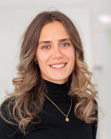 Eine Frau mit langen, lockigen Haaren und einem schwarzen Rollkragenpullover lächelt freundlich in die Kamera. Sie trägt eine goldene Halskette.