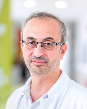 Ein Arzt in einem weißen Kittel mit Brille, der in einem hellen, modernen Raum steht und direkt in die Kamera schaut.