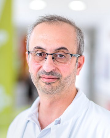 Ein Arzt in einem weißen Kittel mit Brille, der in einem hellen, modernen Raum steht und direkt in die Kamera schaut.