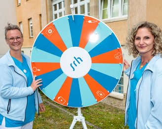 Zwei Frauen in blauen Outfits stehen neben einem Glücksrad mit dem Logo "srh" und verschiedenen Symbolen.
