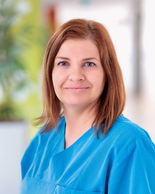 Eine Frau mit langen, braunen Haaren trägt ein hellblaues medizinisches Outfit. Sie steht in einem hellen, modernen Raum und lächelt freundlich.