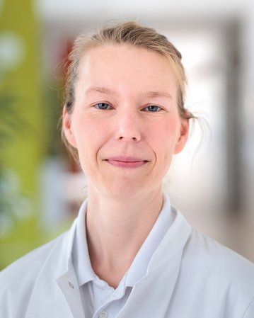 Eine Frau in einem weißen Kittel lächelt freundlich in die Kamera. Der Hintergrund ist unscharf und zeigt eine helle, moderne Umgebung.