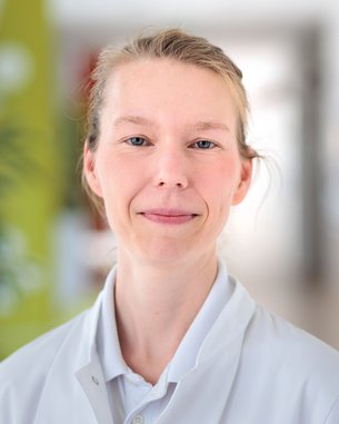 Eine Frau in einem weißen Kittel lächelt freundlich in die Kamera. Der Hintergrund ist unscharf und zeigt eine helle, moderne Umgebung.