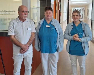 Drei Mitarbeiter in medizinischer Kleidung stehen in einem Flur eines Krankenhauses. Im Hintergrund ist ein Anmeldeschild sichtbar.