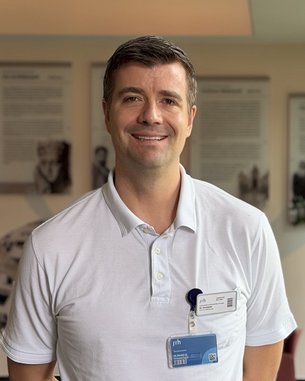 Ein Mann in einem weißen Poloshirt steht in einem modernen Raum mit Pflanzen und Informationswänden im Hintergrund.